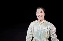 Nahaufnahme der Schauspielerin Miriam Berger in hellem Kostüm vor dunklem Hintergrund, Szene aus „Oinkonomy" am Theater Gütersloh