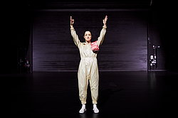 Miriam Berger in hautfarbenem Kostüm steht mit erhobenen Armen auf dunkler Bühne, Szene aus „Oinkonomy" am Theater Gütersloh