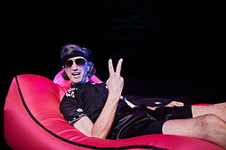Frank Siebenschuh in dunkler Kleidung und Sonnebrille liegt auf einem pinkfarbenen Sofa, Szene aus „Oinkonomy" am Theater Gütersloh