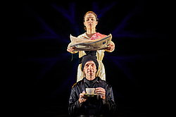 Person in hautfarbenem Kostüm hält eine Zeitung über eine vor ihr hockende dunkel gekleidete Person. die eine Kaffeetasse in der Hand hält, Szene aus „Oinkonomy" am Theater Gütersloh